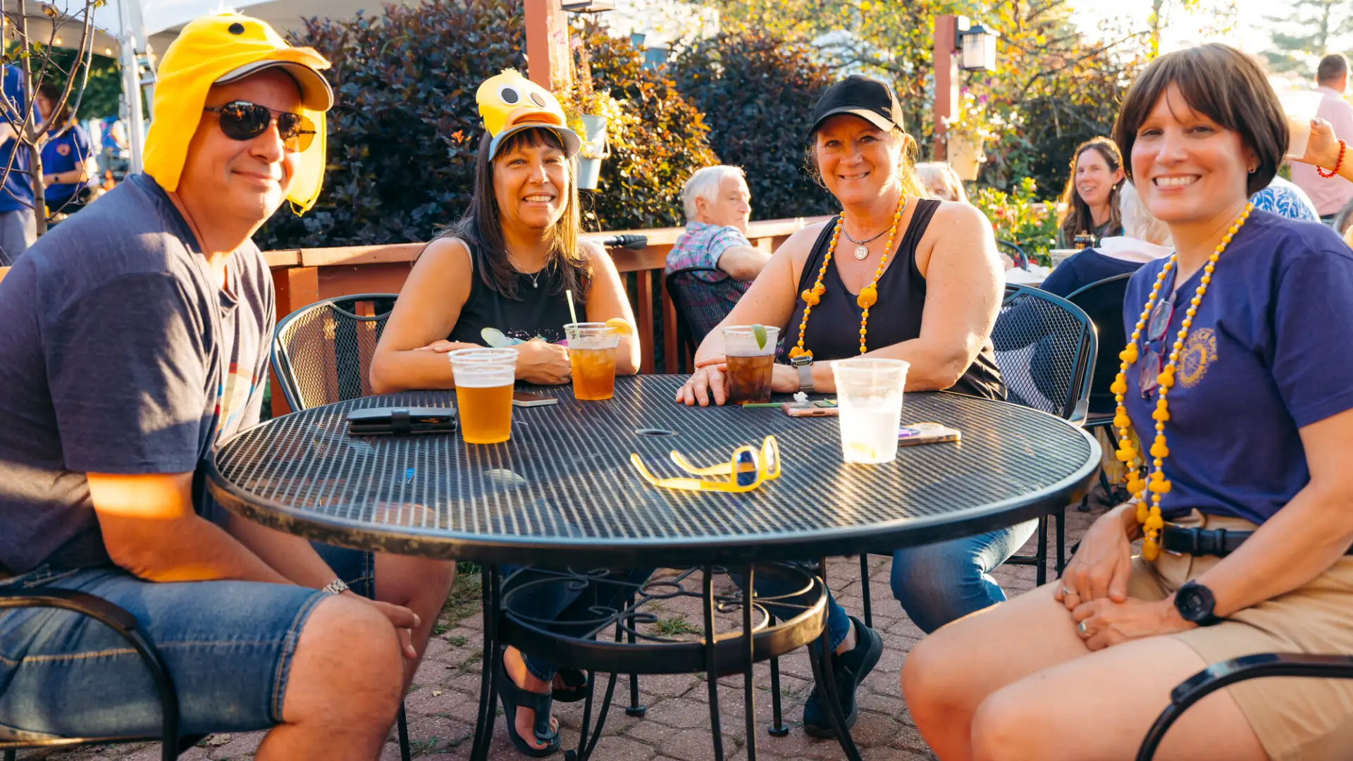 Group of four wearing yellow duck hats, necklaces, and other duck-themed attire sit at a metal deck table during event smiling at camera